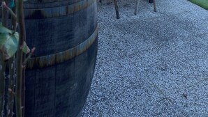 Zona salotto all'aperto con tavolo e sedie in legno, una bottiglia di vino e un bicchiere di vino rosso su una botte.