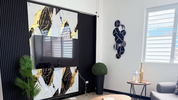 A modern living room with a flat-screen TV mounted on a black and white abstract wall, two round wooden tables, a grey sofa, and a potted plant. - Charming 3BR Near Ponce Attractions (Ponce)