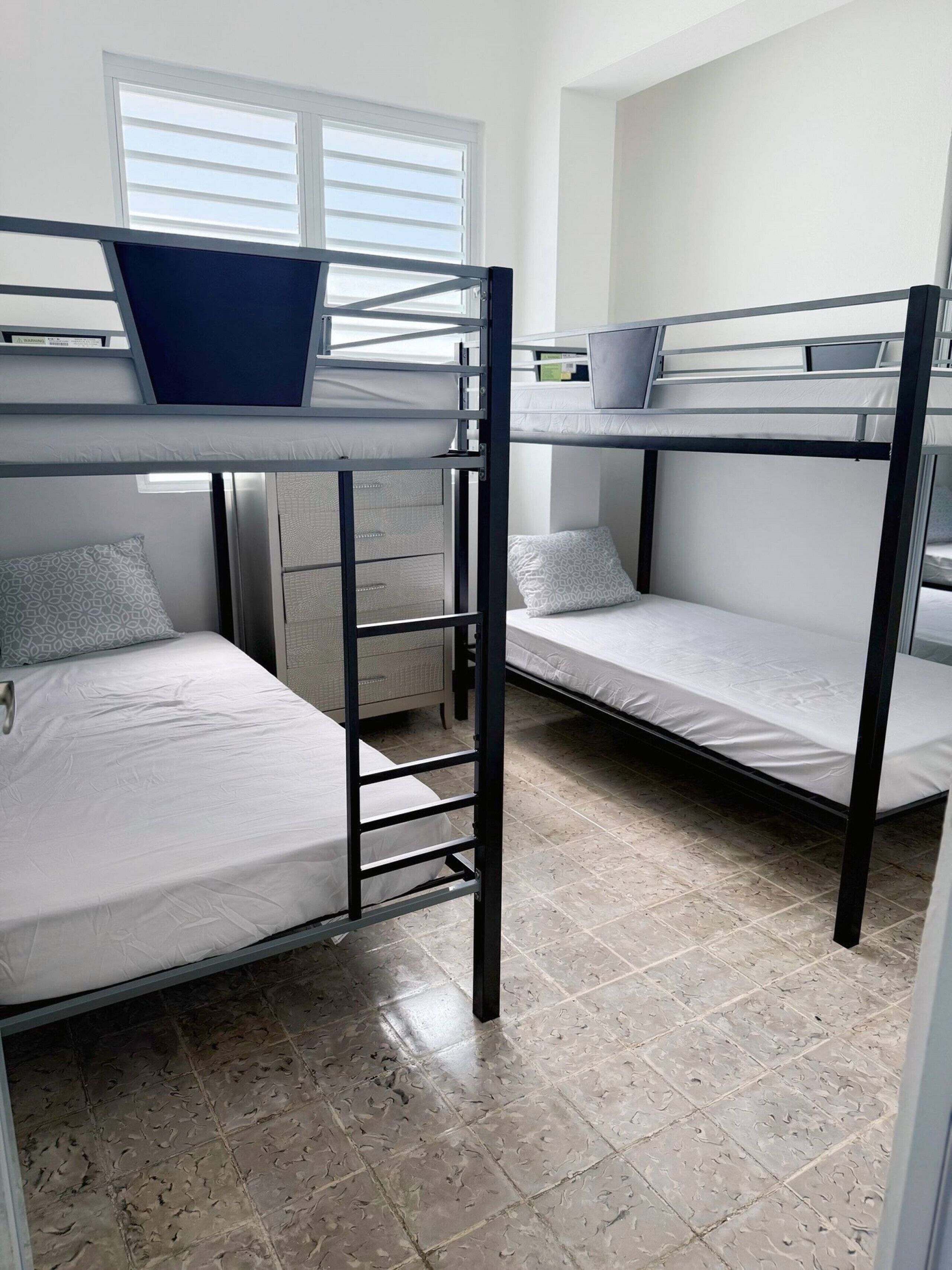 A dormitory room with bunk beds, a desk, and a window with blinds.