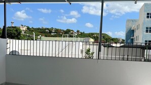 An empty balcony with a metal roof, white walls, and a tiled floor. A fence separates the balcony from a sports field and buildings in the background. - Charming 3BR Near Ponce Attractions (Ponce)