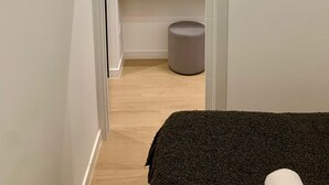Un couloir étroit mène à une pièce où se trouvent un lit, un petit banc et une étagère en bois.