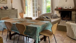 Un coin repas avec une table dressée pour quatre personnes, entourée de chaises en osier. On aperçoit un foyer avec un manteau en pierre et un canapé au fond.