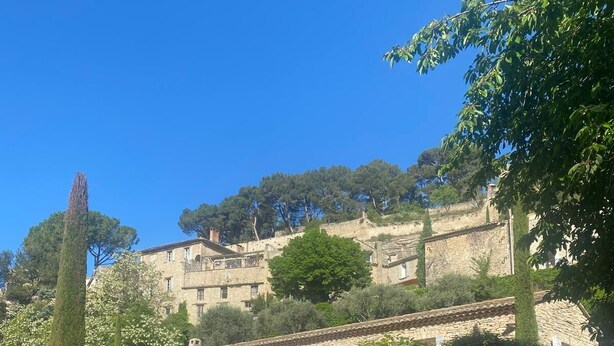 Un bâtiment en pierre doté d’une entrée fermée par un portail, entouré de verdure et surplombé d’un ciel bleu azur.