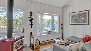 A cozy living room with a red wood-burning stove, a gray sofa, and a large window with a view of trees and a lake. - Sunrise Bay Cottage - Your Perfect Escape on Stoney Lake, 1.5 hrs from the GTA (Trent Lakes)