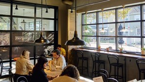 Een café met grote ramen, houten balken in het plafond en hangende lampen.