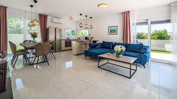 Ein modernes Wohnzimmer mit einem blauen Sofa, einem Essbereich mit Stühlen, einer Küche mit Kühlschrank und großen Fenstern mit Blick auf Grünflächen.