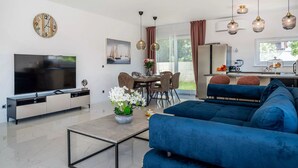 Ein modernes Wohnzimmer mit Flachbildfernseher, einem blauen Sofa, einem Essbereich mit Tisch und Stühlen sowie einer Küchenzeile mit Kühlschrank und Kaffeemaschine.