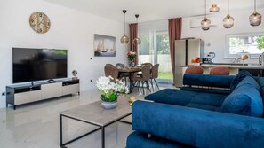 A modern living room with a flat-screen TV, a blue sofa, a dining area with a table and chairs, and a kitchen area with a refrigerator and a coffee machine.