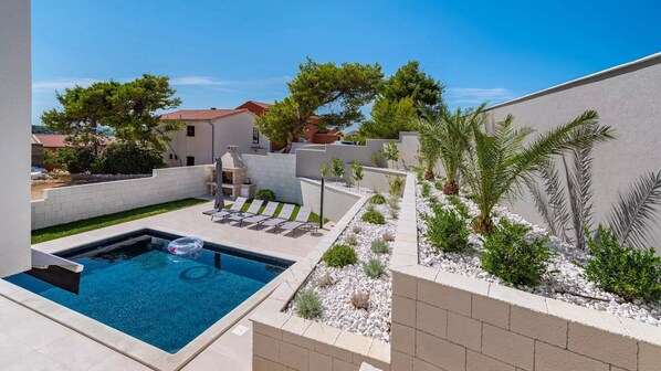 A swimming pool with lounge chairs and a barbecue area, surrounded by a landscaped garden with palm trees and shrubs.