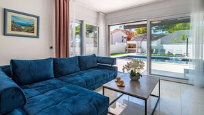 Ein modernes Wohnzimmer mit einem blauen Sofa, einem Glastisch und Blick auf einen Swimmingpool sowie eine Sitzgelegenheit im Freien.