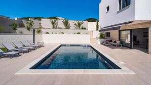 Ein rechteckiger Pool mit Fliesen umgeben, mit Liegestühlen und einer kleinen Sitzecke inklusive Sofa und Couchtisch.