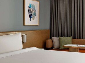 A hotel room with a bed, a wooden headboard, a nightstand, a table, and a couch with pillows. - By Charles River | Luxe Stay. Fitness + Dining (Boston)