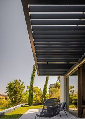 A modern outdoor patio with a pergola, a black wicker chair, and a view of a landscaped garden. - Villa GA A Luxury 3BR With Sauna and Pool (Ishëm)