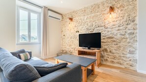 A modern living room with a stone wall, a flat-screen TV, a grey sofa, and a wooden coffee table. - Holiday Home 'La halte provençale - Cocooning' with Private Terrace, Wi-Fi & Air Conditioning (Châteaurenard)