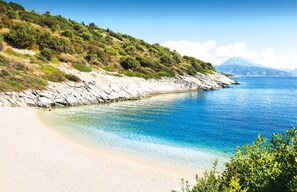 Eine Küstenlandschaft mit sandigem Strand, klarem blauen Wasser, felsigen Klippen und einem gebirgigen Hintergrund.
