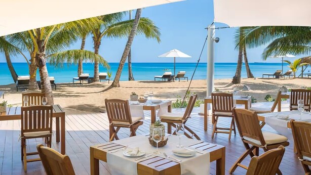 Un restaurante frente a la playa con terraza al aire libre, vistas al océano, palmeras y sillas de playa.