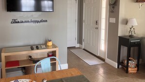 Ein Wohnzimmer mit Fernseher, Schreibtisch, Stuhl, Esstisch, einer Tür, einer Lampe und einer Wanduhr.
