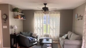 Ein Wohnzimmer mit einer Couch, einem Couchtisch, einer Lampe, einem Deckenventilator und einem Fenster mit gemusterten Vorhängen.