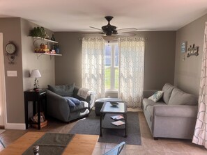 A living room with a sofa, a coffee table, a lamp, a ceiling fan, and a window with patterned curtains.