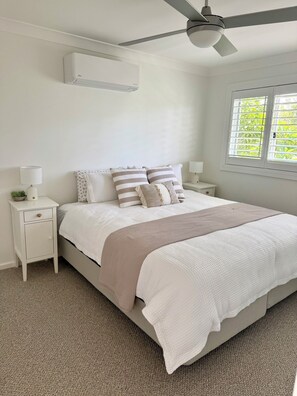 Une chambre à coucher comprenant un lit, des tables de chevet, un ventilateur de plafond, un appareil de climatisation et une fenêtre avec des stores.