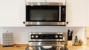 Een keuken met een ingebouwde magnetron, oven en kookplaat. Er zijn witte kastjes, een houten aanrechtblad en een groene ovenhandschoen hangt aan de muur.