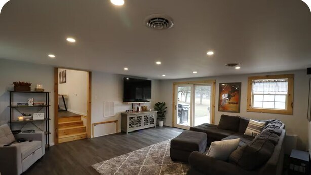 Een ruime woonkamer met een donker houten vloer, een grote modulaire bank, een salontafel en een aan de muur bevestigde televisie. Een trap leidt naar een ander niveau, en een groot raam zorgt voor veel natuurlijk licht.