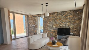 A modern living room with a stone wall, a sofa, a coffee table, a TV, and a large window. - EL-LU 2 Villa with breathtaking 360-degree seaview and a private pool (Kamilari)