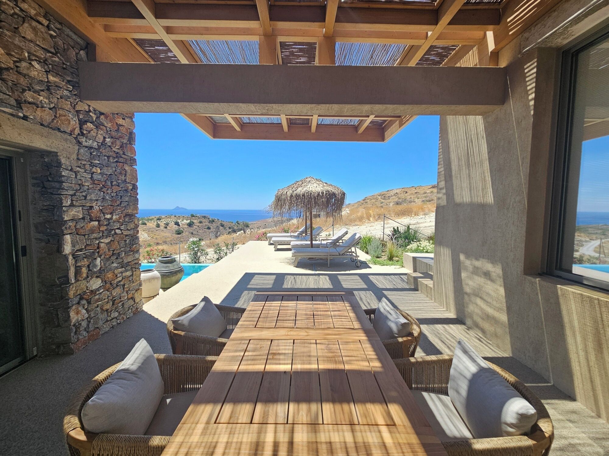 An outdoor seating area with a view of the sea, stone walls, and a thatched umbrella.