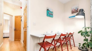 Un coin repas avec une table blanche, des chaises rouges et une lampe noire. Il y a des tableaux au mur et une plante sur le sol.
