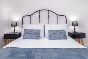 A neatly made bed with a white and blue patterned bedspread, flanked by two black nightstands with lamps and drawers. - The Snug by the Sea - Central Whitby, Beach Front (West Cliff)