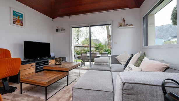 Een moderne woonkamer met een flatscreen televisie, een houten salontafel, een grijze bank en een eetgedeelte zichtbaar door glazen deuren.