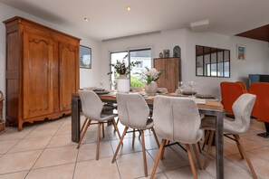 Uma sala de jantar com um armário de madeira, cadeiras estofadas cinzentas, uma mesa de madeira e uma janela com vista para casas.