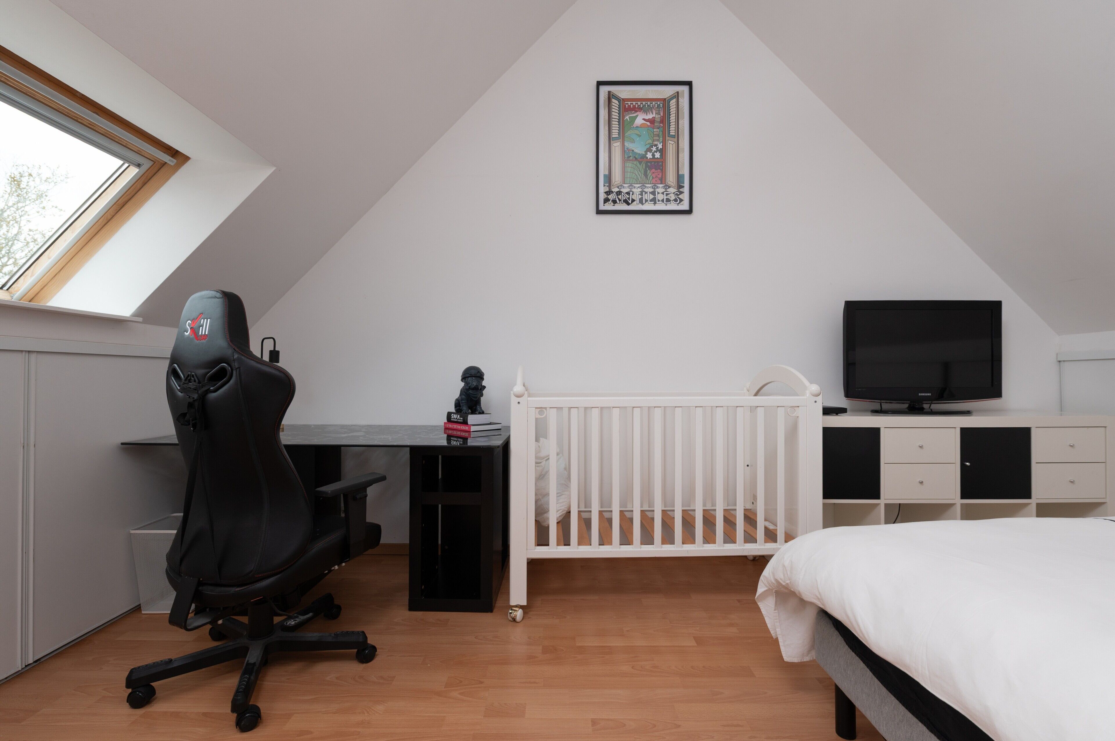 Un dormitorio con techo inclinado, un escritorio con computadora, un televisor, una cuna y una ventana.