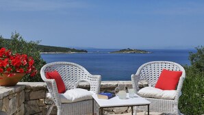 Zwei Korbstühle mit roten Kissen, ein Tisch mit einem Buch und einer Kanne, und Blick auf das Meer und Inseln.