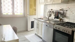 A kitchen with white cabinets, a gas stove, a dishwasher, and a window with curtains. - Apartment in a quiet street (Schiltigheim)