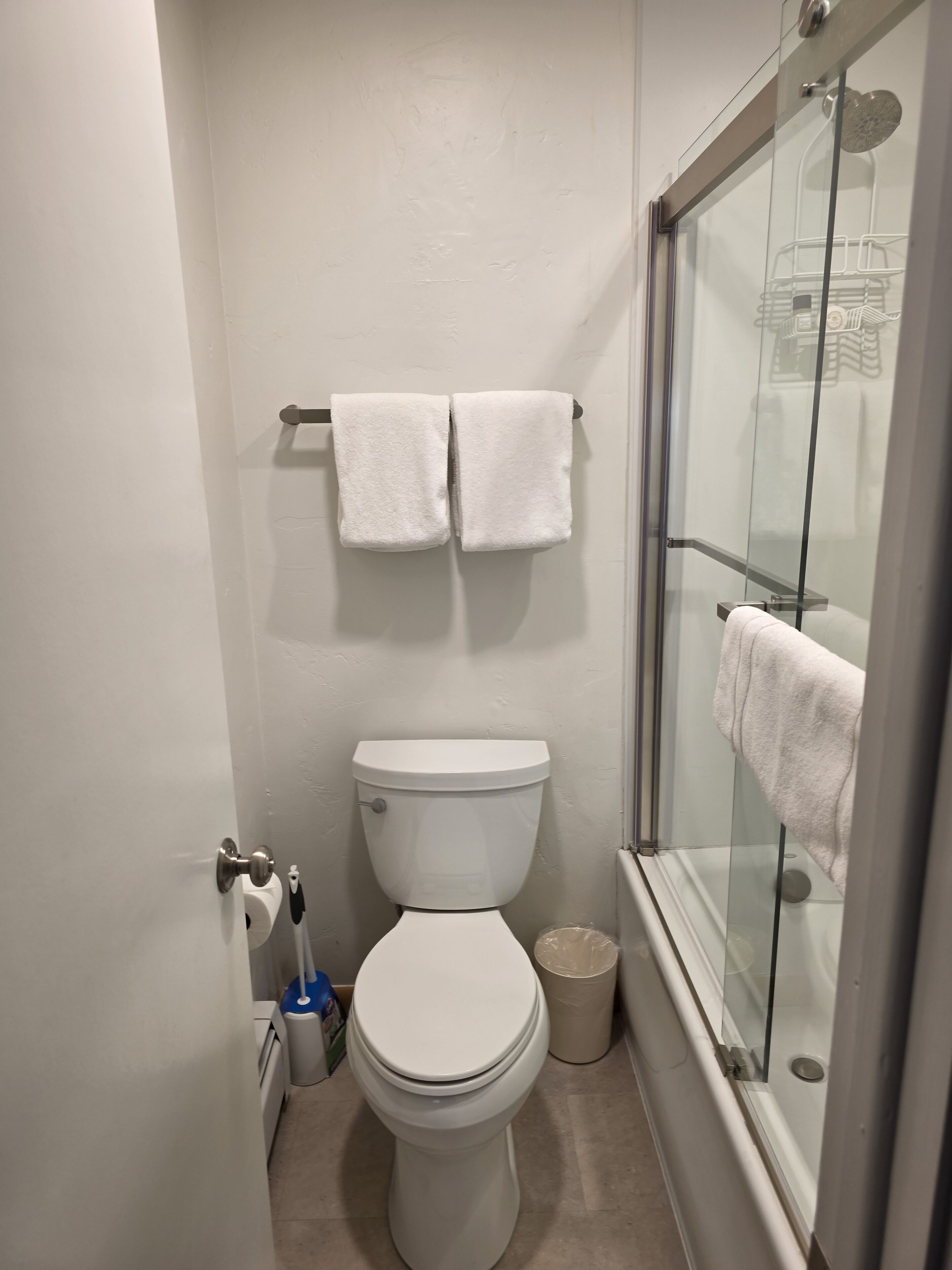 A small, clean bathroom with a toilet, two towels on a rack, a glass shower enclosure, and a trash can.