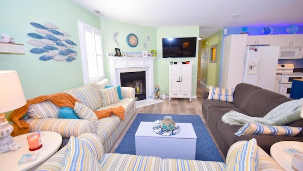 A living room with striped sofas, a fireplace, a television, and a kitchen area with cabinets and appliances. - Montego Bay-colonial Drive 13339 3 Bedroom Home (Ocean City)