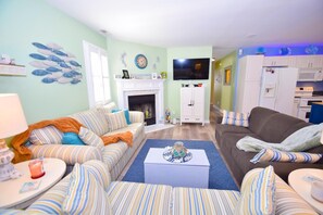 A living room with striped sofas, a fireplace, a television, and a kitchen area with cabinets and appliances. - Montego Bay-colonial Drive 13339 3 Bedroom Home (Ocean City)