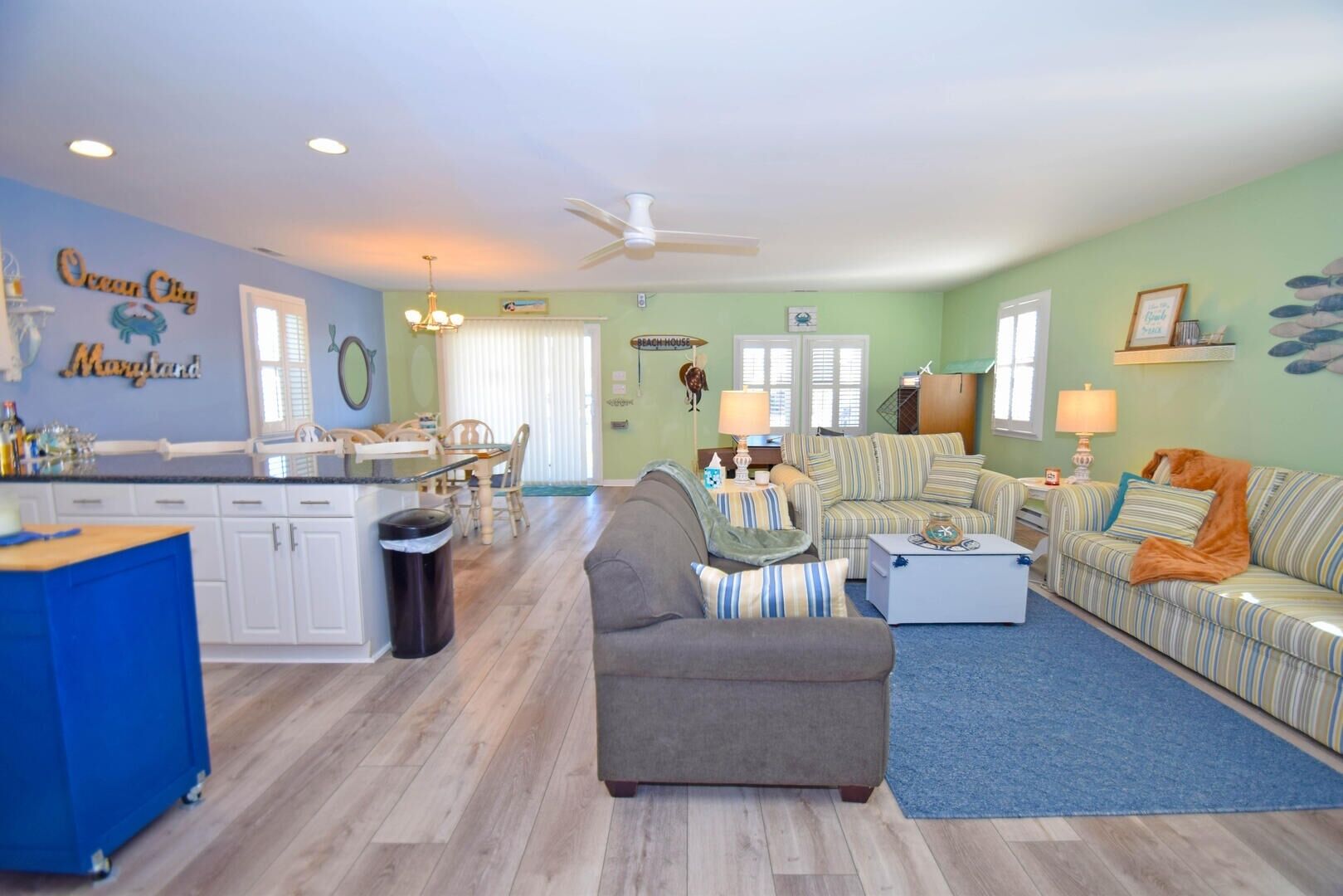 A spacious living room with a gray sofa, striped couches, and a blue rug. A kitchen with white cabinets and a blue countertop is visible.