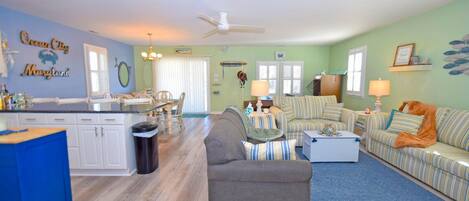 A spacious living room with a gray sofa, striped couches, and a blue rug. A kitchen with white cabinets and a blue countertop is visible.