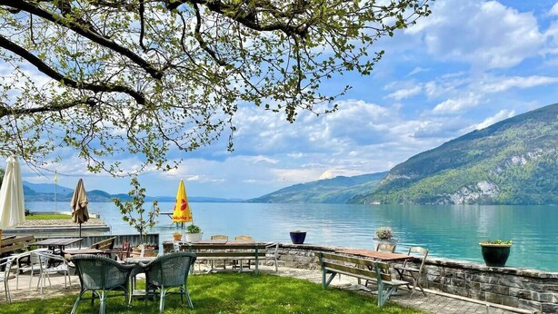 Ein Sitzbereich im Freien mit Tischen und Stühlen mit Blick auf einen See, umgeben von Grünflächen und Steinmauern.