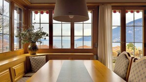 Ein Essbereich mit einem großen Tisch, hölzernen Bänken, einer Blumenvase und Bergblick durch große Fenster.