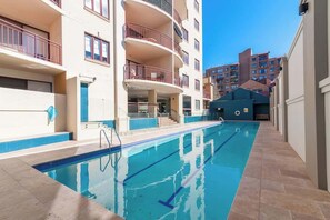 Une piscine à l’eau d’un bleu cristallin, entourée d’immeubles à plusieurs étages dotés de balcons, sous un ciel dégagé.