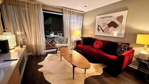 Una sala de estar moderna con un sofá rojo, una mesa de centro de madera y un tapiz de cuero de vaca. Hay un ventanal con cortinas, un cuadro en la pared y una lámpara sobre la mesita de noche.