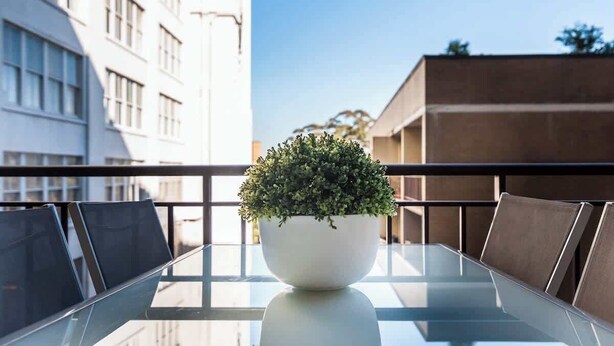 Una mesa con cubierta de vidrio, con un cuenco blanco que contiene una planta, rodeada de sillas, en un balcón con barandilla.