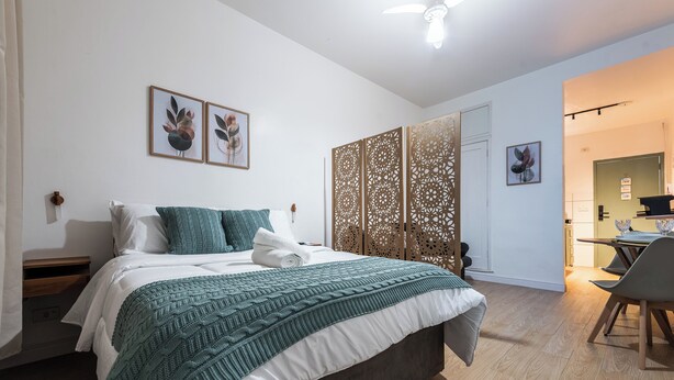 A bedroom with a bed, pillows, a blanket, a wooden floor, a partition, and a desk with chairs. - StayHome 1min da Praia | São Vicente SP | Família ou Casal (São Vicente)