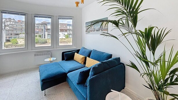 A modern living room with a blue sectional sofa, a small round table, and a large potted plant. - Casa Vento -  Koksijde (Koksijde)