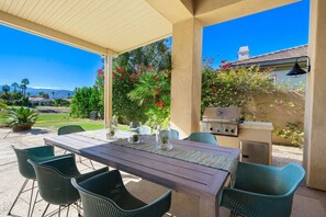 An outdoor patio with a wooden table and chairs, a grill, and a view of a residential area with greenery and a clear sky. - 4 Bedroom Private Pool Home in Mira Vista (Rancho Mirage)