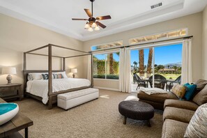 A bedroom with a four-poster bed, a sitting area with a sofa and ottoman, a ceiling fan, and a view of a landscaped garden through large windows. - 4 Bedroom Private Pool Home in Mira Vista (Rancho Mirage)