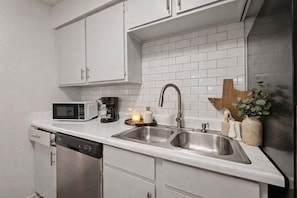 A kitchen with white cabinets, a double sink, a microwave, a coffee maker, and a small plant. - Entertainment District Escape • 2BR + Parking (Arlington)